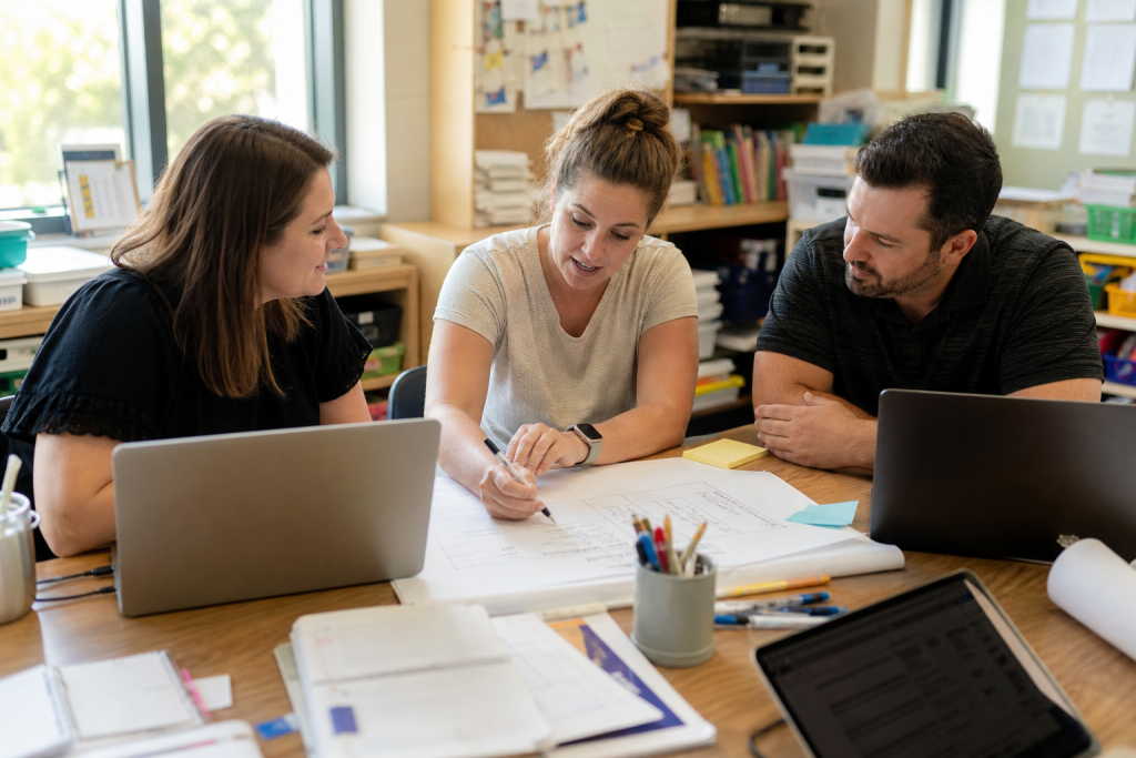 Small team of educators plans shared adoption around one table with laptops, notes, and a practical rollout discussion.
